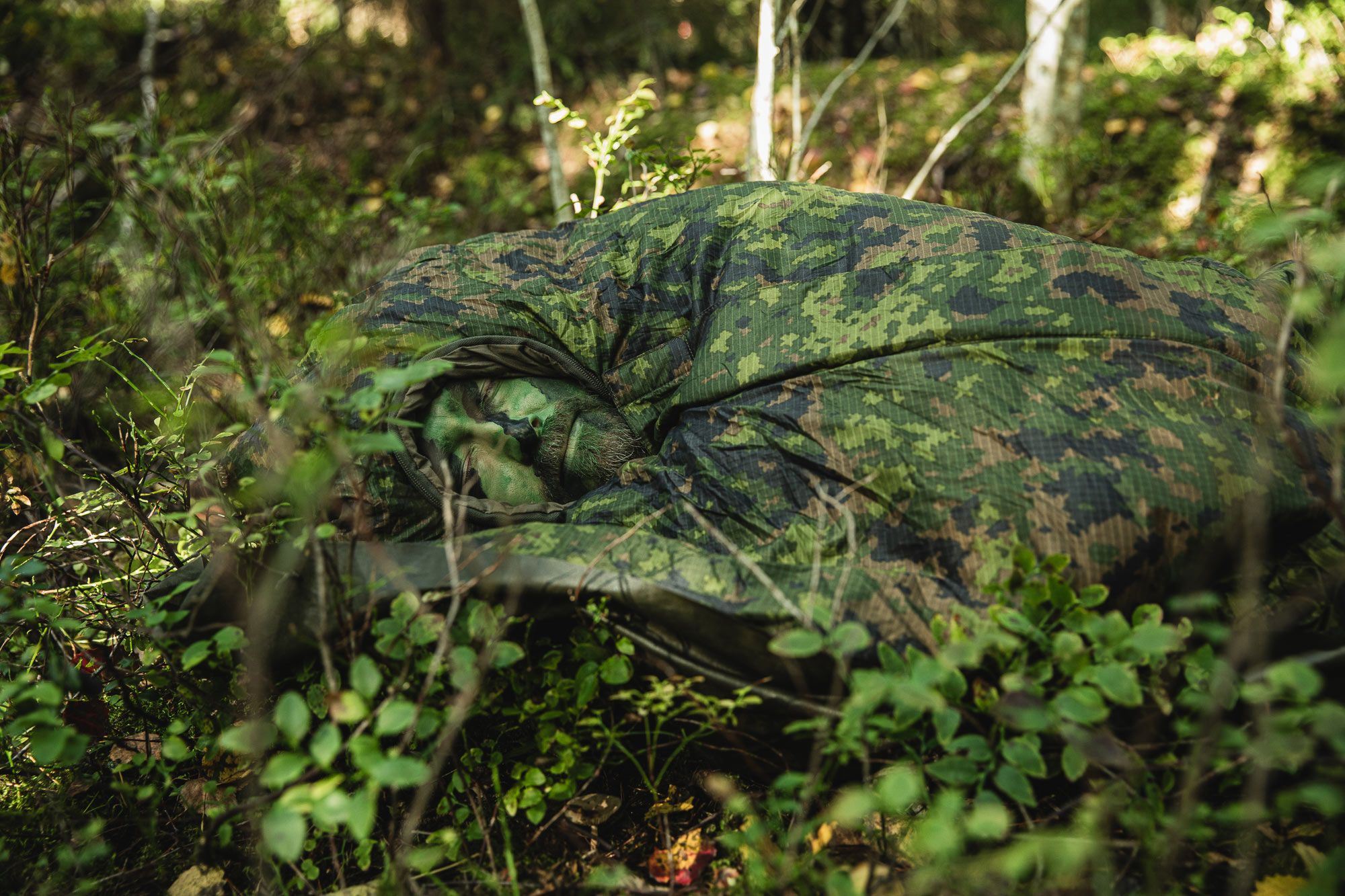 Mies makaa M05-kuvioidussa makuupussissa metsässä.