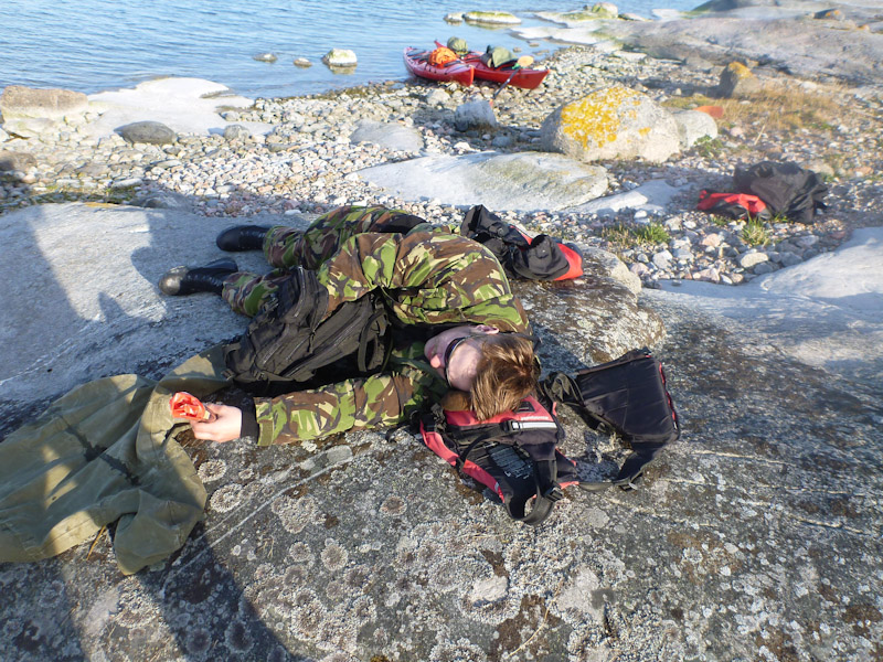 Mies makaa rantakalliolla pelastusliivit tyynynään ja käsi repun sisällä.