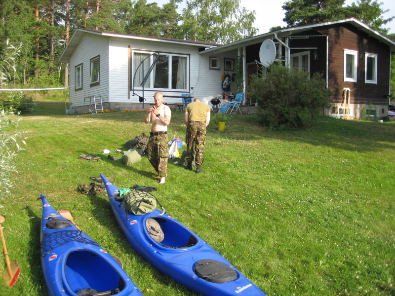 Kaksi miestä ja kaksi sinistä kanoottia sekä varusteita omakotitalon pihalla.