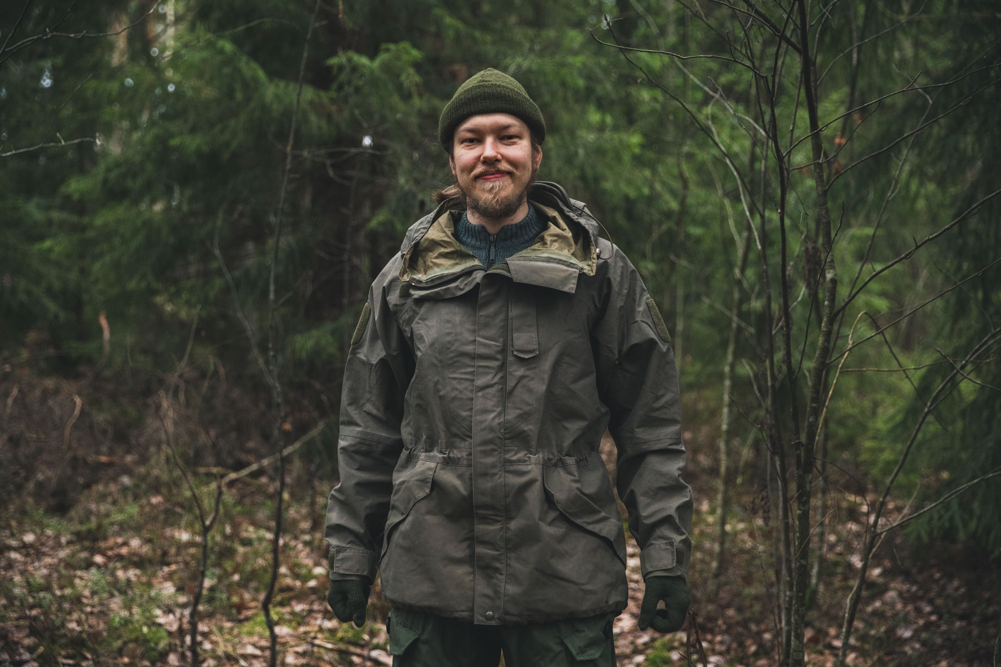 Hymyilevä mies metsässä päällää harmaanvihreä takki.