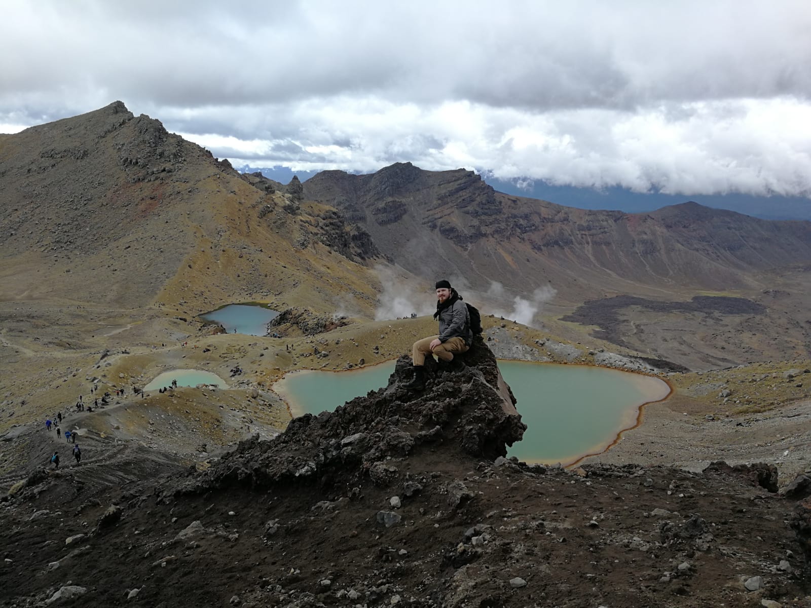 Mies istuu kukkulan päällä, taustalla vuoria ja turkoosin värisiä vuoristojärviä.