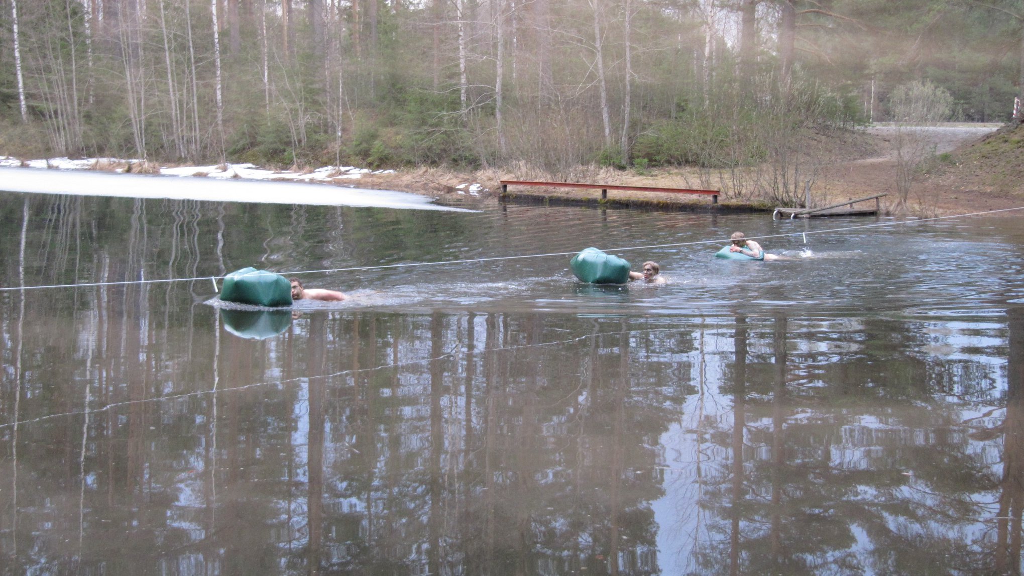 Kolme miestä ylittää vettä uiden mukanaan vihreät kelluvat säkit. Vesistö on rannasta jäässä.