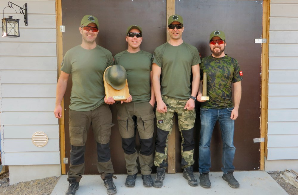 Neljä miestä poseeraa ovien edessä hymyillen. Vasemman puoleisilla on käsissään plakaatti, johon on kiinnitetty sotilaskypärä.