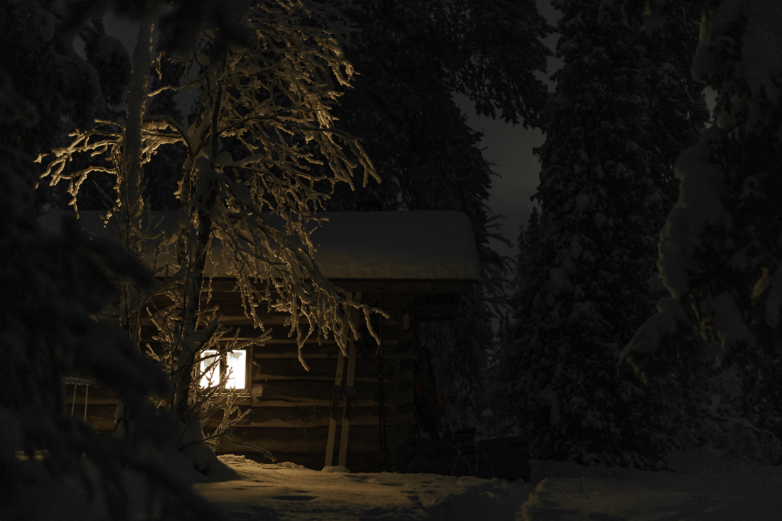 Tupa, jonka ikkunasta paistaa valo, pimeässä lumisessa metsässä.