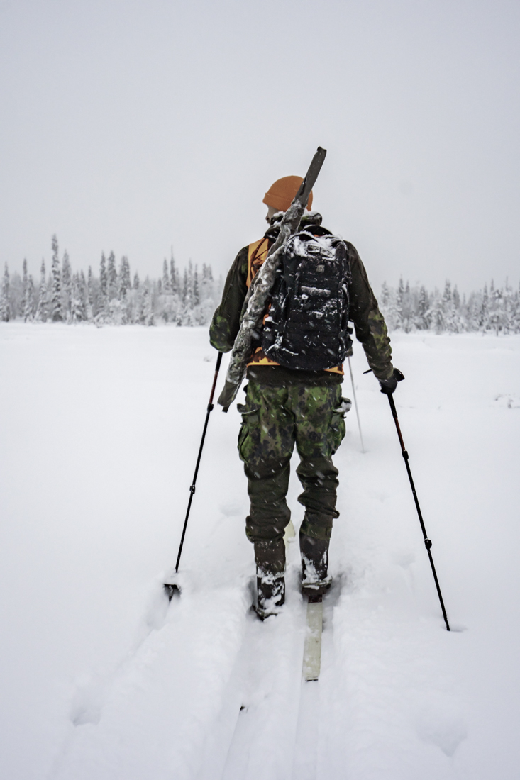 Metsästäjä hiihtämässä harmaassa talvikelissä aukion poikki.