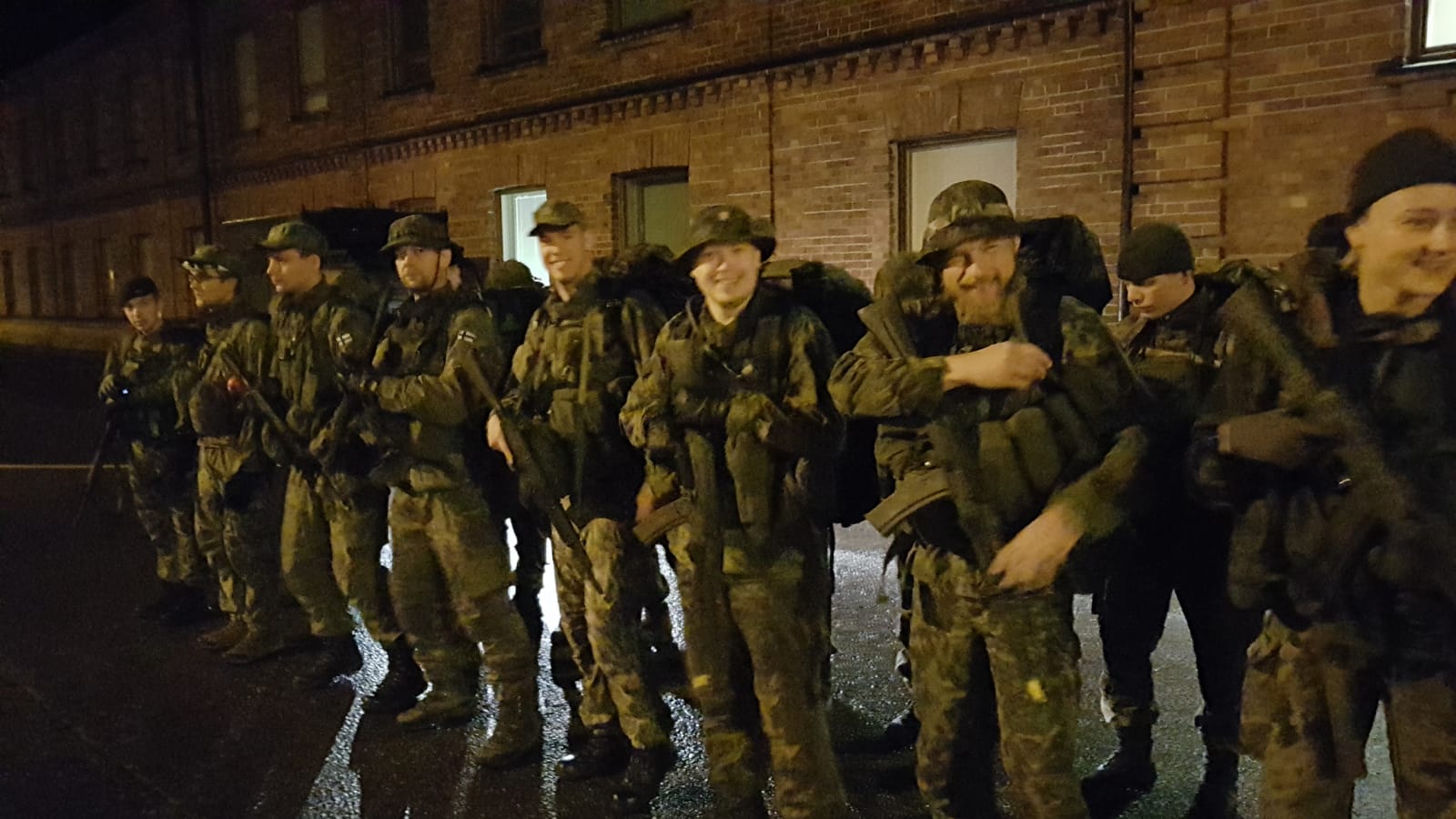 Hymyileviä sotilaita hämärässä rivissä tiilirakennuksen edessä.