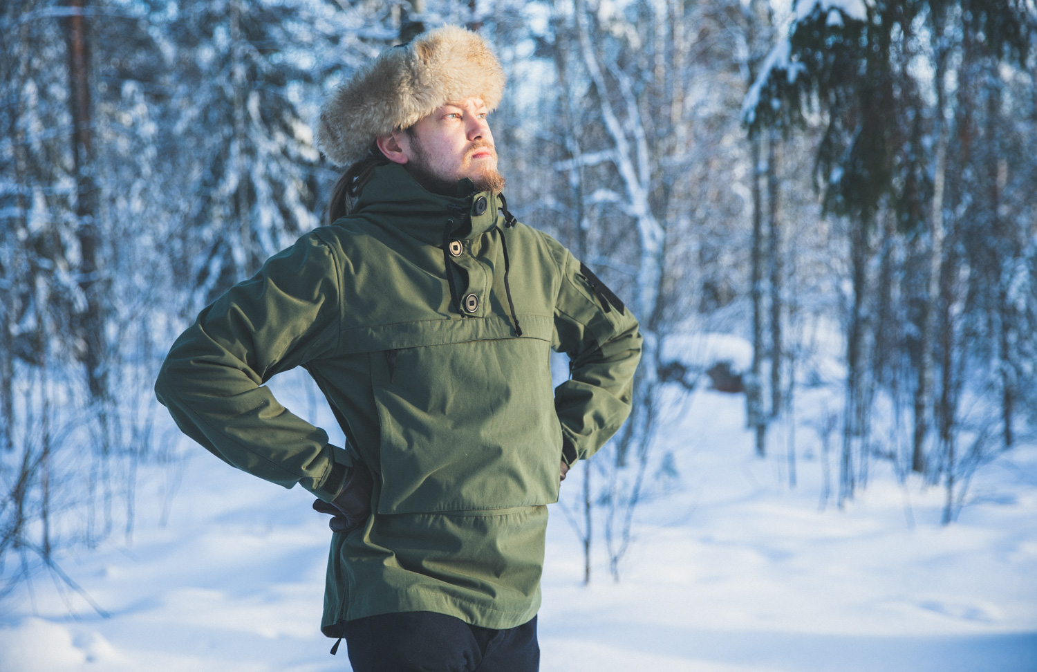 Mies pukeutuneena karvalakkiin, anorakkiin ja kylmän kelin vaatteisiin lumisessa metsämaisemassa.