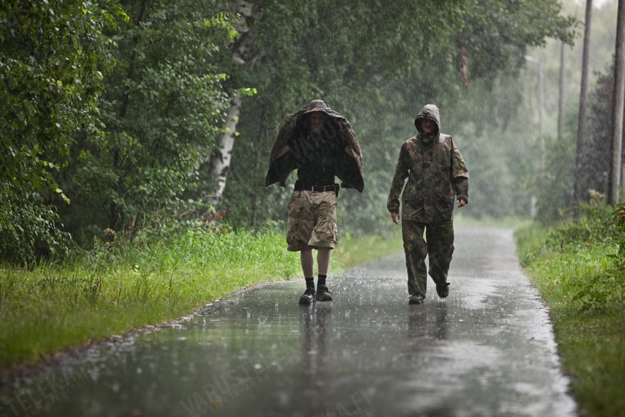 kaksi henkilöä kävelee sateessa. Toisella on päällään sadevaatteet, toisella reisitaskushortsit.