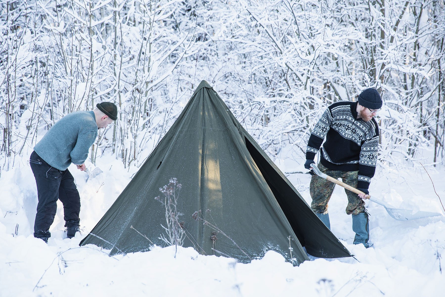 Two guys at a snowy forest edge on either side of a tent set up in the snow, showeling snow from around the tent.
