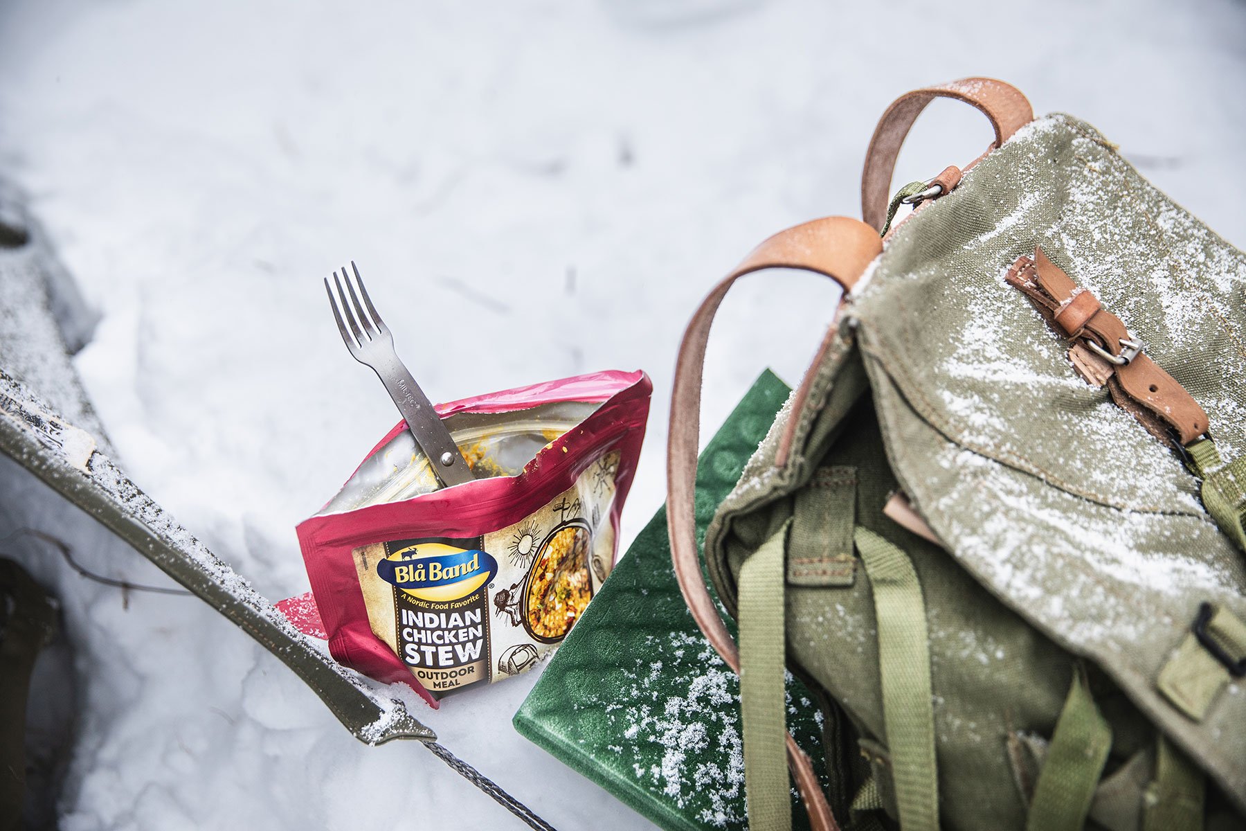 A Blå Band outdoor meal bag in the snow with a fork sticking out of it, next to a seating mat and a backpack.