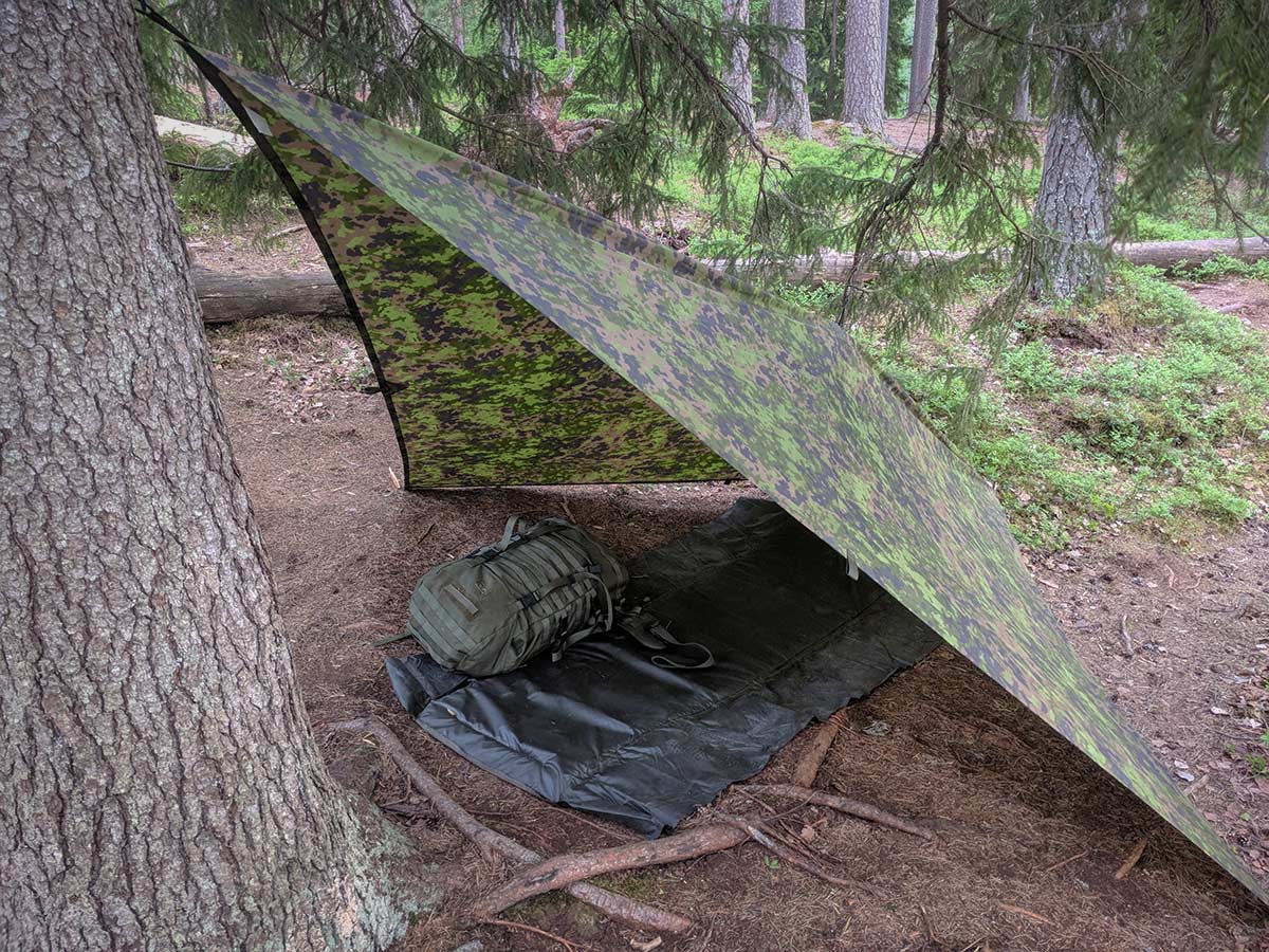 Savotta FDF Sleeping Pad spread out under a camouflage-patterned tarp in a forest.