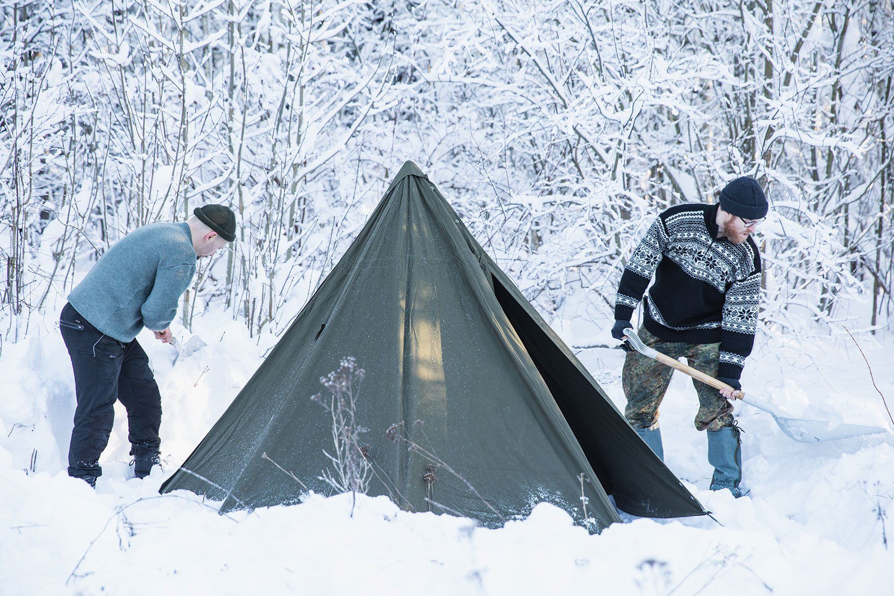 Teltta pystytettynä lumeen lumisen metsän reunaan. Kaksi miestä lapioi lunta teltan ympäriltä.