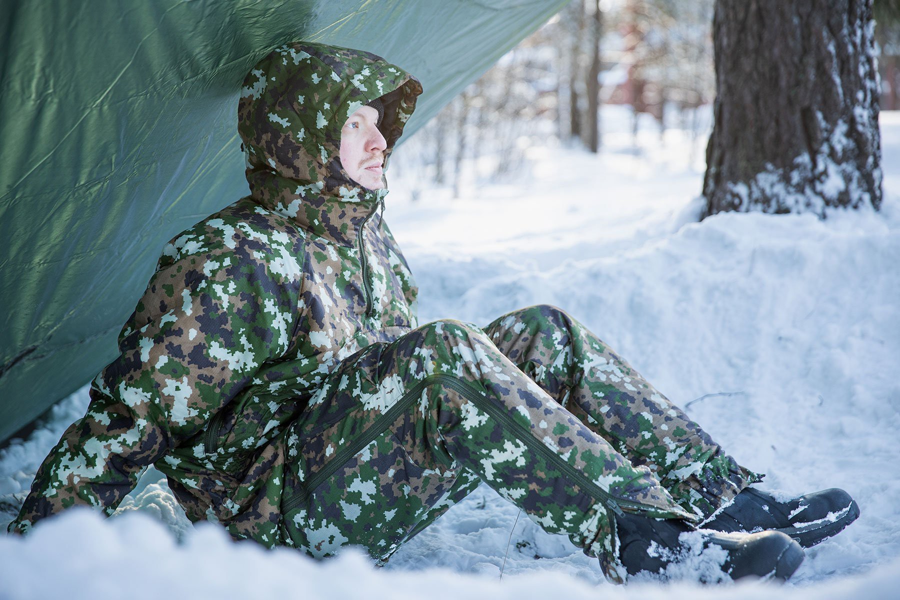 Mies istuu laavukankaan alla lumihangessa maastokuvioiduissa talvivaatteissa